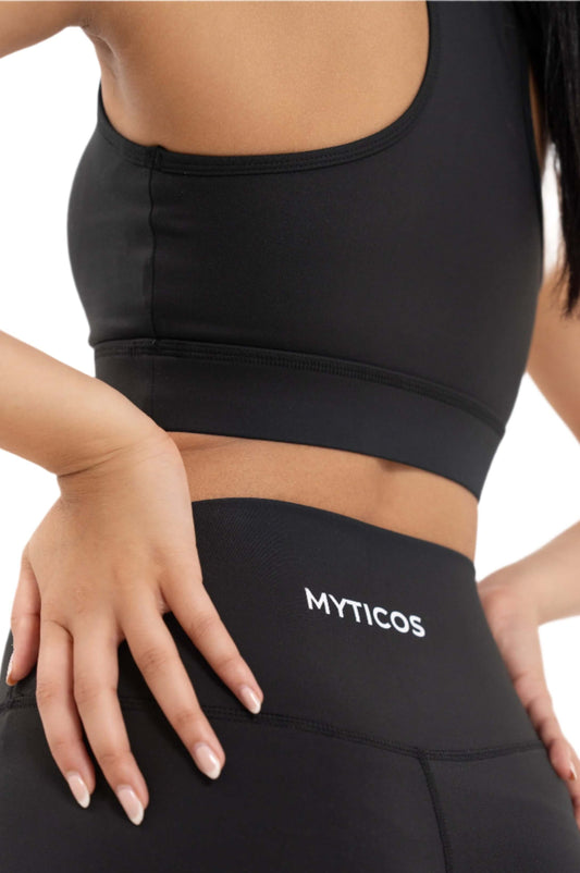 Close-up view of Myticos®️ logo on the waistband of black women's leggings, highlighting the fabric texture, stitching, and snug fit against a white background.