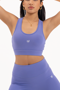 Woman posing with arms raised wearing Myticos®️ lavender sports bra and leggings set, highlighting activewear fit and flexibility against a white background.